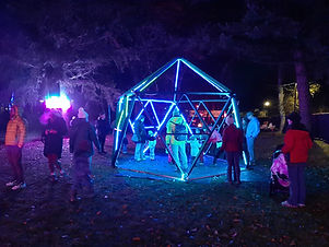 A wooden geodesic dome with people surrounding it and neon rope lights illuminated